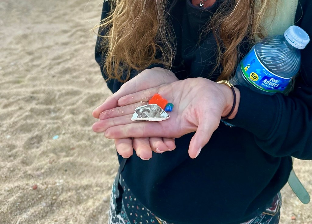 Orange sea glass with a piece of pottery in a woman's hand who is also holding a plastic water bottle on a beach in Barcelona, Spain