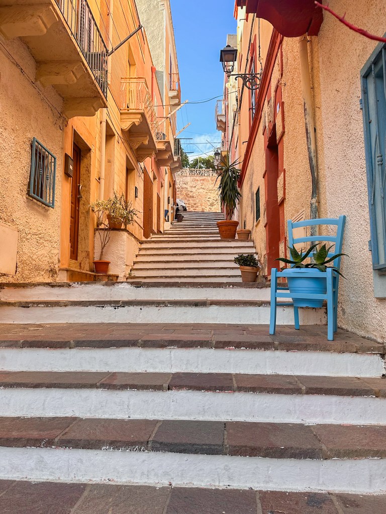 A brick staircase in the middle of orange buildings leading up to another street with a blue chair holding a potted plant outside of a window. 