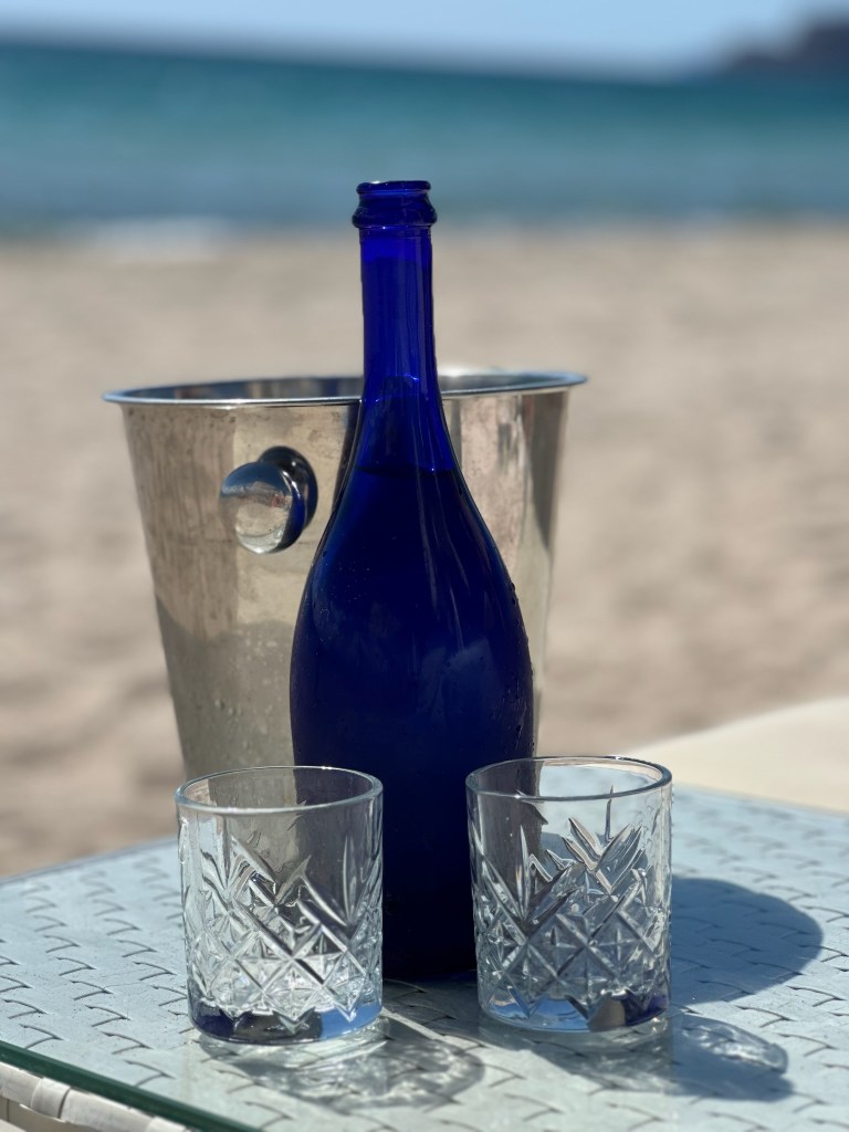 A blue glass bottle of water with two crystal drinking glasses on a table on the beach in Sardinia, Italy
