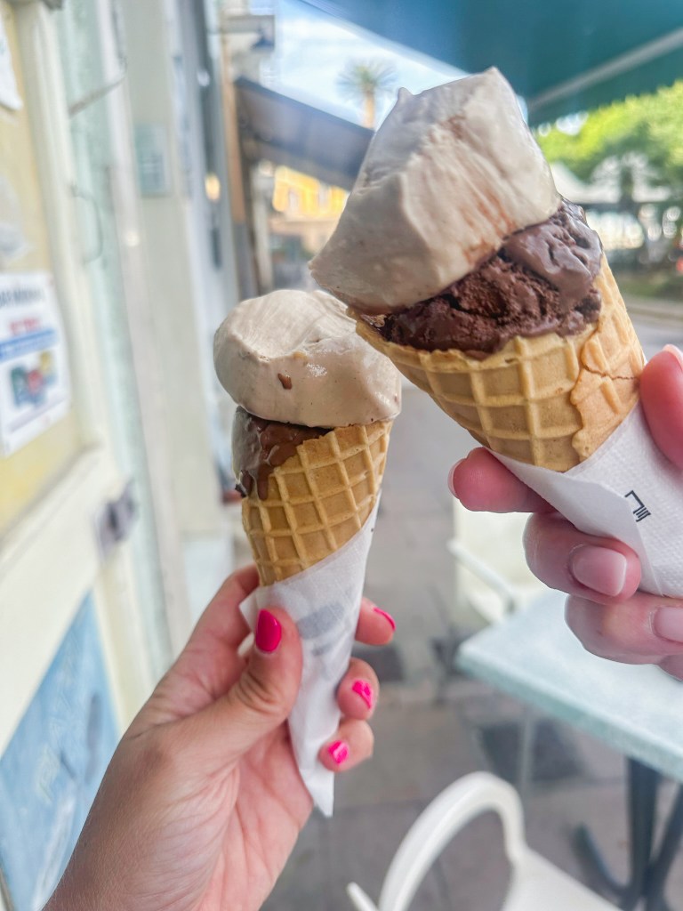 Cheers-ing a gelato cone with a friend, nails painted pink with picnic tables in the background in Carloforte, an island of San Pietro, Italy. 