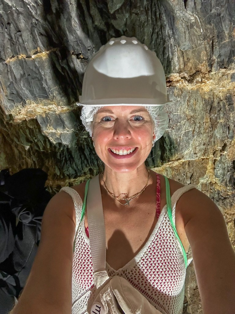 Jules, a travel blogger, mother and pilot wife, wearing a bathing suit coverup, Lululemon crossbody and a hard hat in the Masua mine tunnel