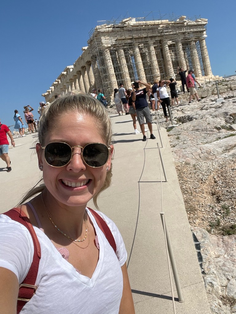 Jules, pilot wife and blogger standing in front of the acropolis in Athens, Greece, wearing a white t-shirt and sunglasses