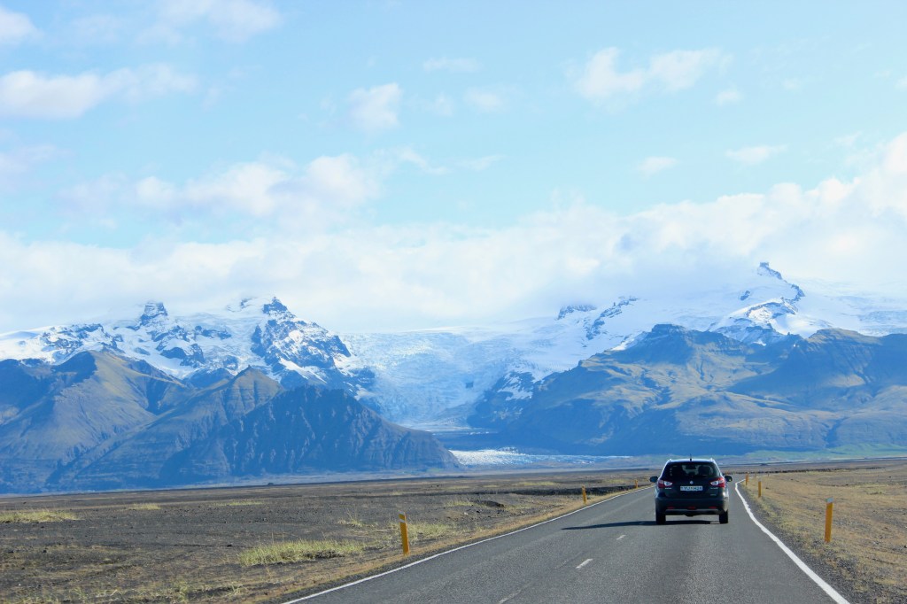 scenic picture from a car in Iceland with another car ahead with flat land to the side and glaciers ahead