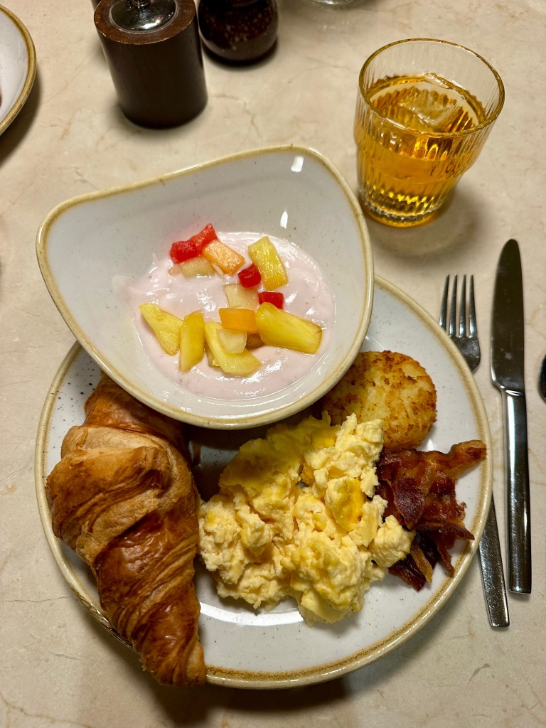 Plate of eggs, croissant, bacon and hasbrowns, with a bowl of yogurt and a glass of orange juice for a Amsterdam Hotel Breakfast