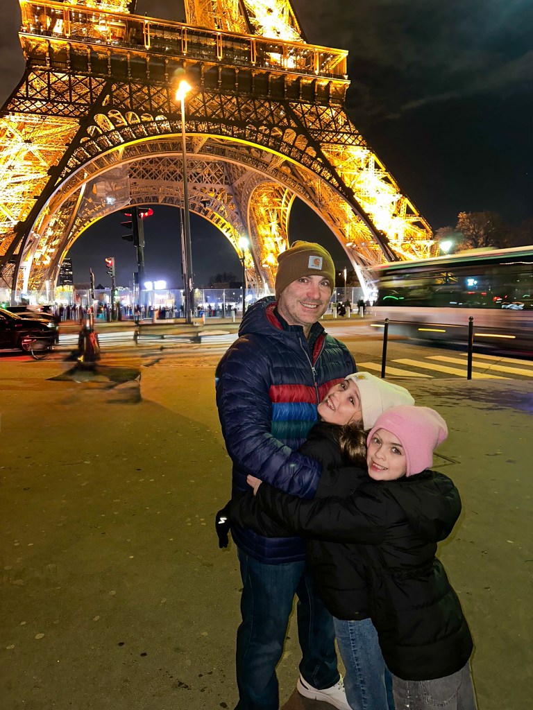Father and daughters posed in front of the base of the Eiffel Tower at night 