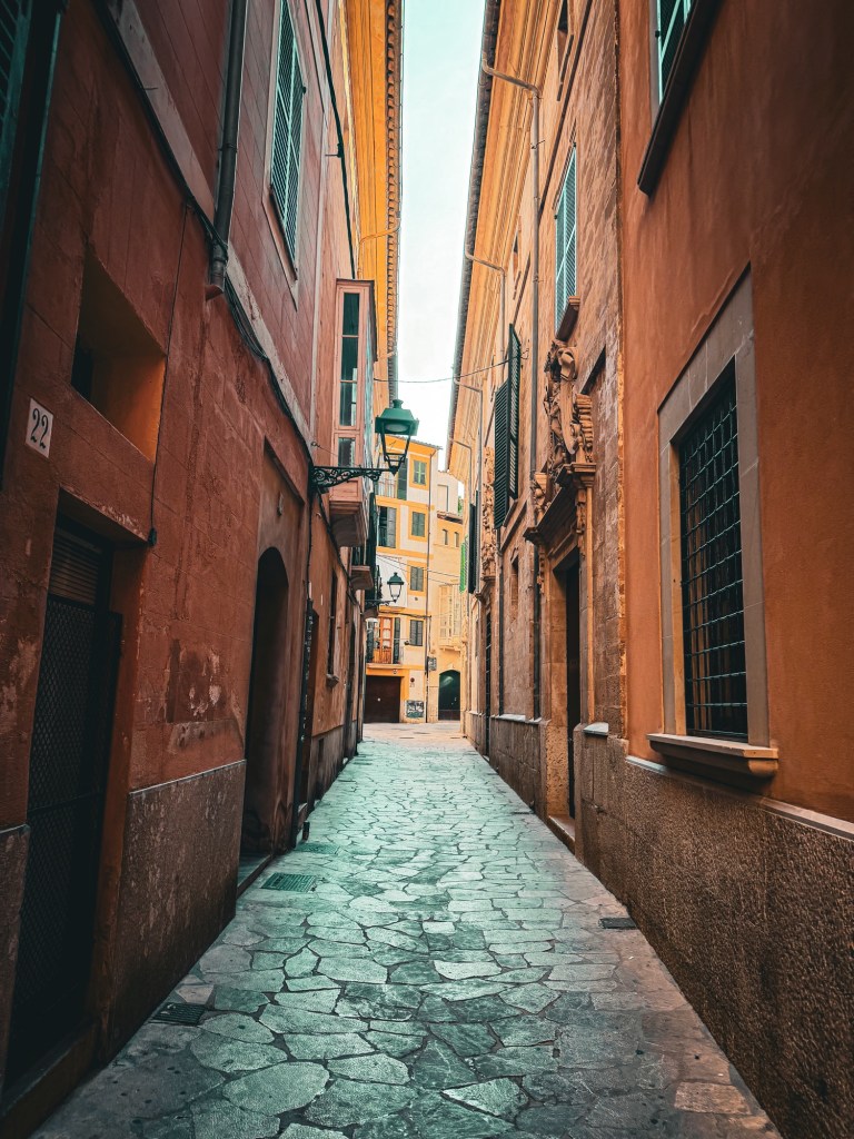 A narrow cobblestone street in the old town of Palma de Mallorca, Spain with tall rust colored buildings on each side of the street.